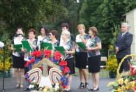 grupa kobiet na scenie w podobnych strojach oraz zielonymi teczkami w dłoniach